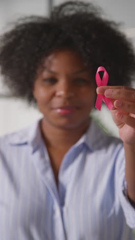 vertical video close up of smiling mid adult woman in hospital clinic holding pink breast cancer awareness ribbon and putting it on her shirt