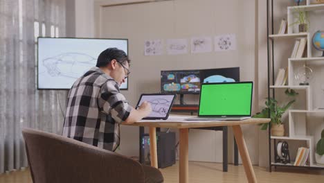 side view of asian male having a backache while drawing car concept on tablet beside green screen laptop in the studio with tv and computers display 3d electric car model