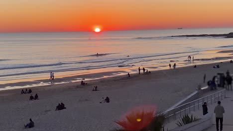 vista amplia y cinematográfica de la gente en la playa de carcavelos, caminando durante la marea baja sobre la arena en el hermoso océano atlántico, puesta de sol de color amarillo intenso sobre reflejos en el agua, portugal