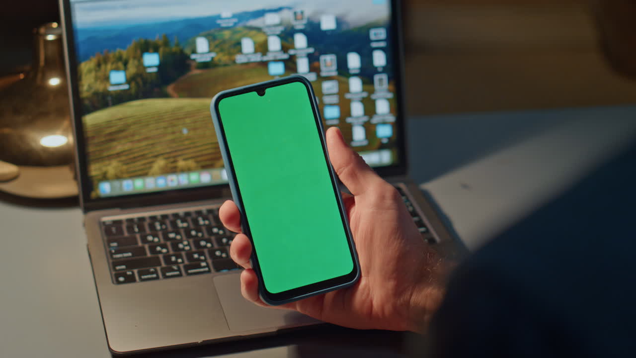 Unrecognizable guy scrolling telephone green screen on laptop workplace closeup
