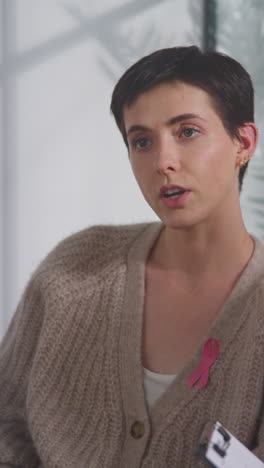 video vertical de una mujer joven con una cinta rosa de concienciación sobre el cáncer de mama reuniéndose y hablando en un grupo de apoyo terapéutico para pacientes con tratamiento de cáncer con un consejero 1
