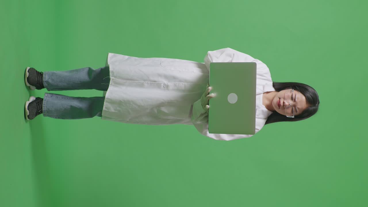 cuerpo lleno de mujer científica asiática que tiene dolor de cabeza mientras está de pie y trabajando en una computadora portátil en el laboratorio de fondo de pantalla verde