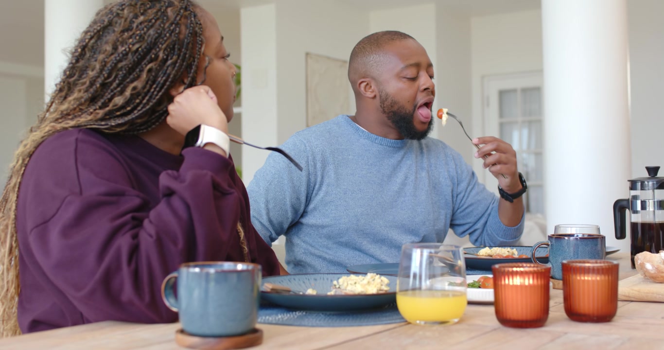 Couple sitting at dining table, woman removing glasses while man eating eggs and looking, talking