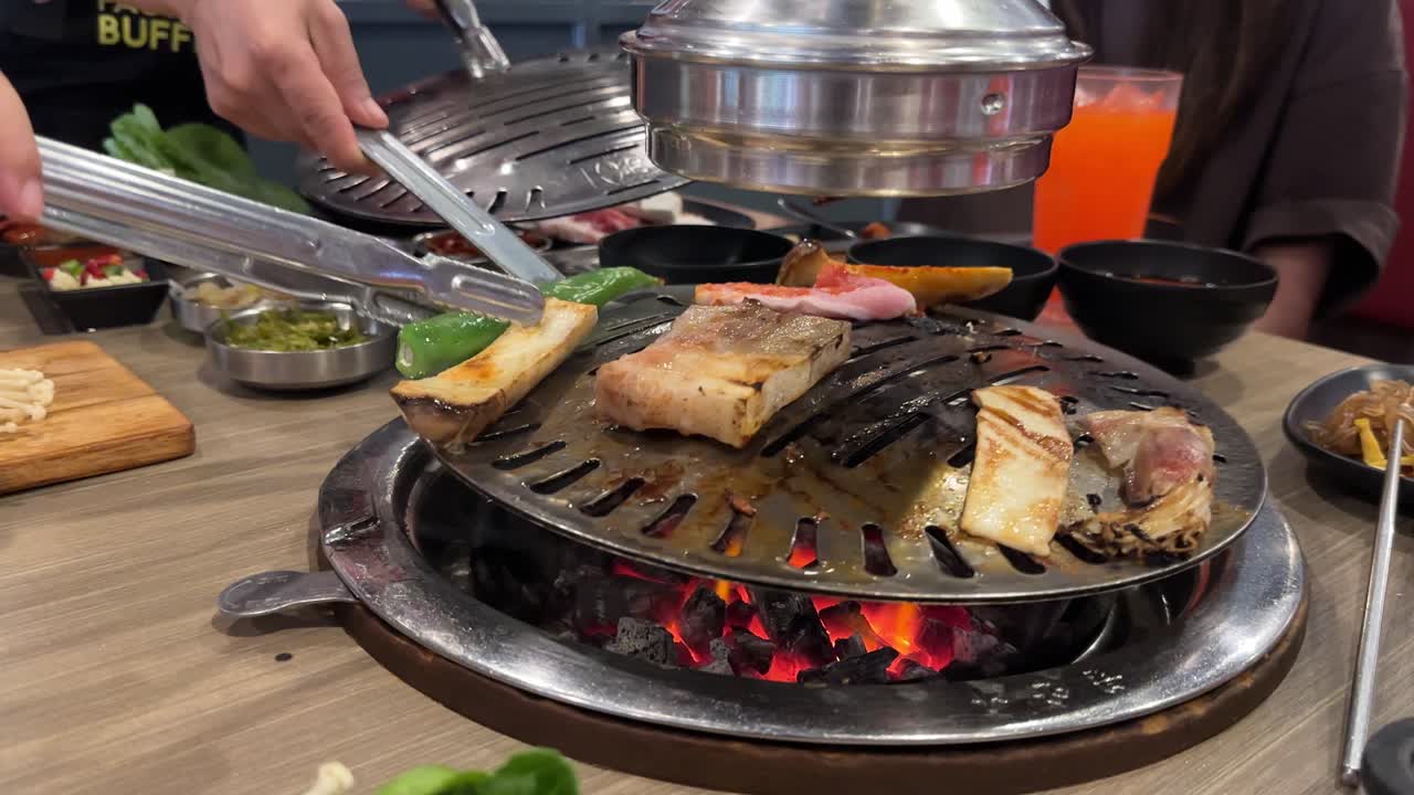 Hands grill pork and vegetables on a tabletop BBQ in a lively Bangkok restaurant. Vibrant atmosphere with colorful dishes and drinks