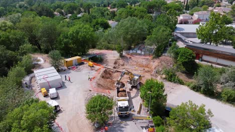 aerial-drone-view-of-a-building-site-surrounded-by-trees,-with-a-backhoe-picking-up-the-soil-to-put-it-on-a-truck,-workers-on-the-site,-safety-barriers