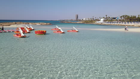 Low-Aerial-of-Pedal-boats-moored-in-turquoise-shallow-water-of-Nissi-Beach,-Ayia-Napa-Cyprus