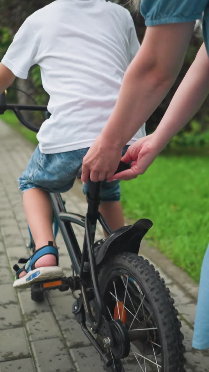 una mujer sostiene el asiento de la bicicleta de su hijo, ayudándole a mantener el equilibrio mientras viajan a lo largo de una pasarela pavimentada rodeada de vegetación