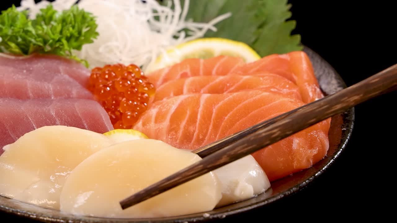 Chopsticks delicately pick up a scallop from a vibrant sashimi sushi set with salmon, tuna, roe, and garnish, under bright studio lighting and black background