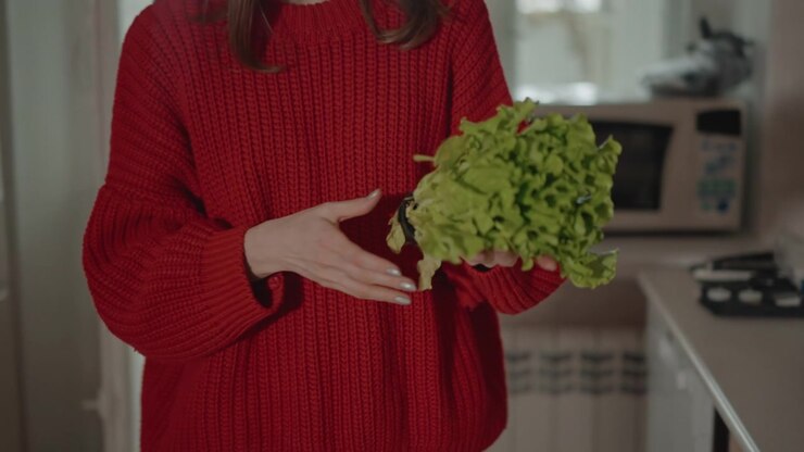 Young woman in red knitted sweater moves through small apartment kitchen, opening light door, entering pantry space, reaching refrigerator shelf, and arranging fresh produce on counter