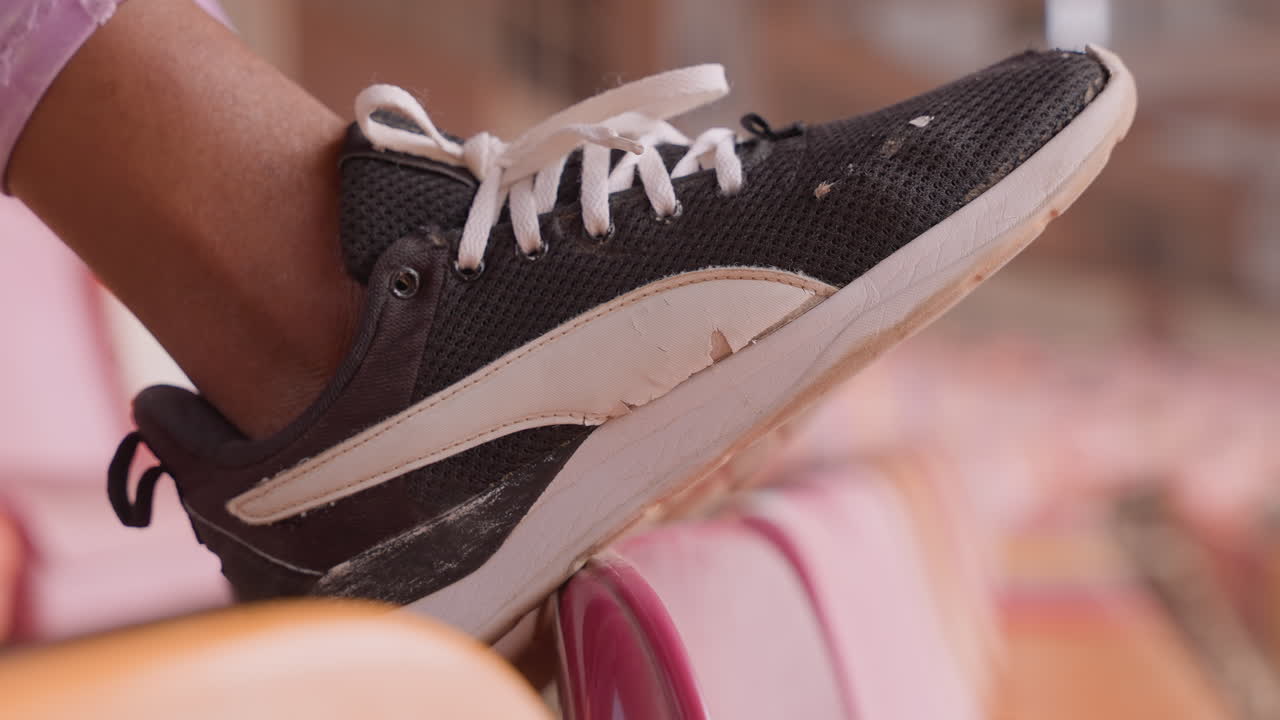 Piede di colore in sneaker sul sedile, adolescente casual che si riposa tra una partita e l'altra, lacci allentati, tomaia in tessuto a maglia testurizzato e intersuola polverosa, illuminazione calda e soffusa, profondità di campo ridotta sfoca lo sfondo dello stadio.