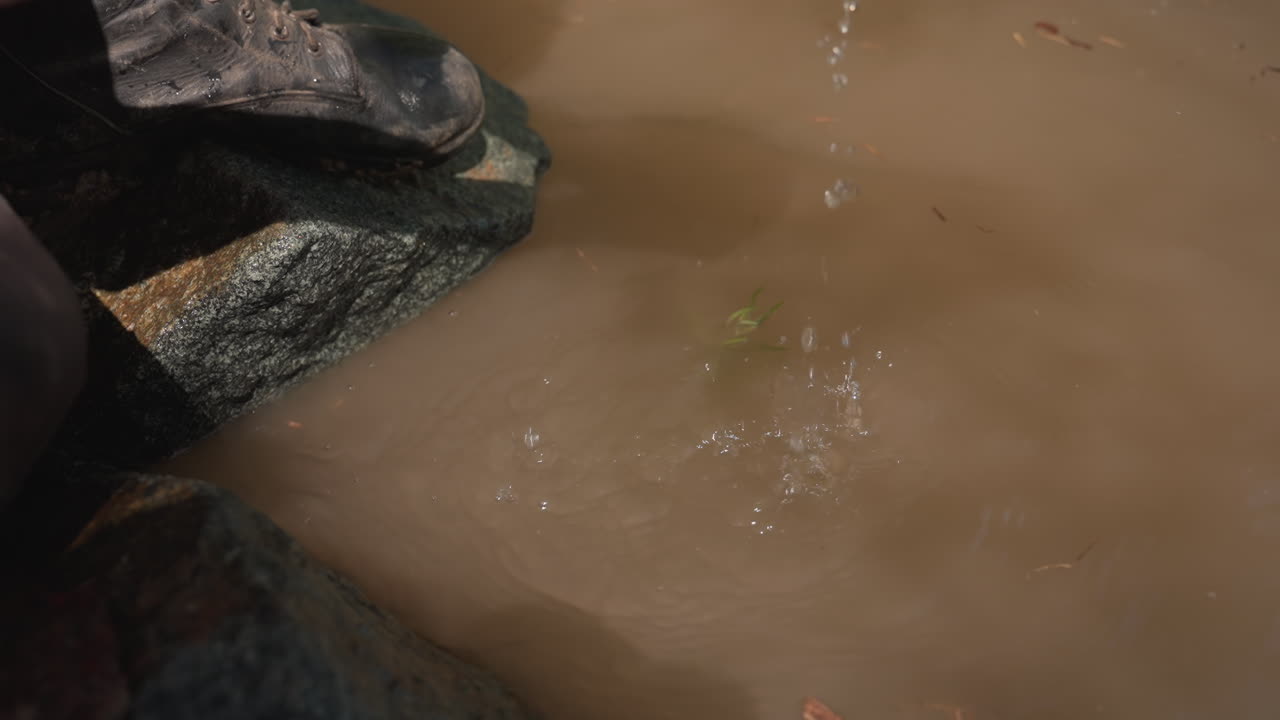 shirtless warrior scoops muddy river water with palms and splashes gently against mossy stones on rugged river bank under bright sunlight and breezy foliage in tranquil outdoor scene
