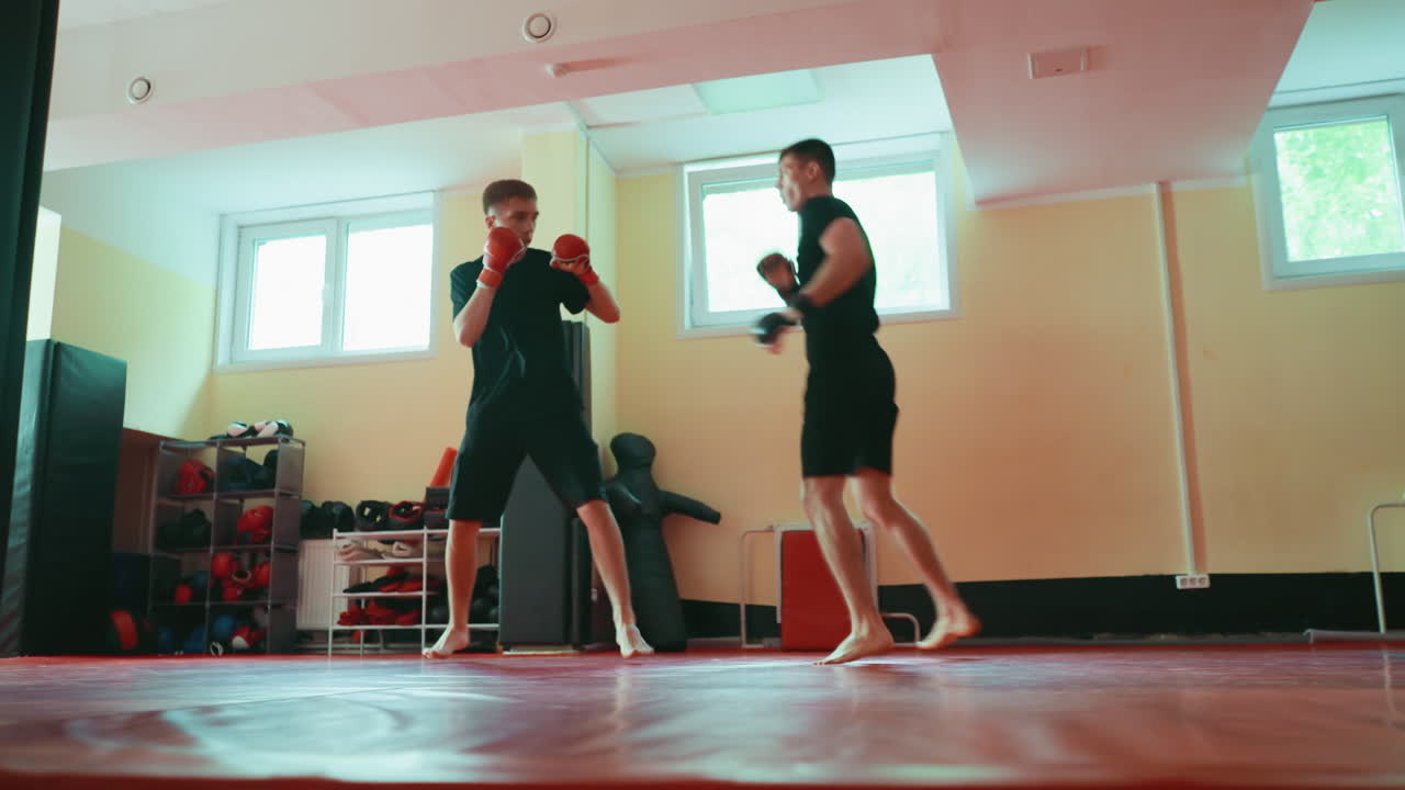 Two fighters sparring in gym on red mat, barefoot, wearing gloves, practicing martial arts combat stance, preparing to exchange punches,strength, and fighting performance under bright natural light
