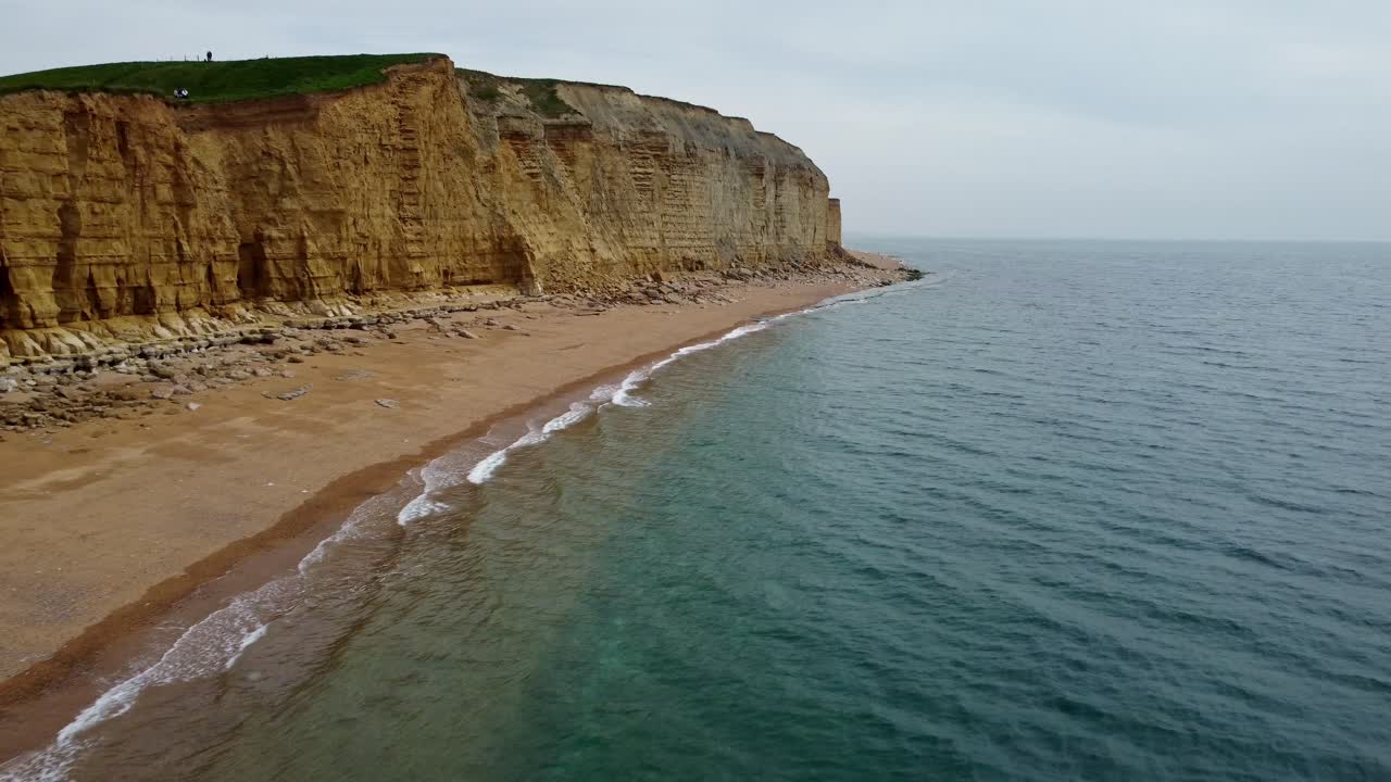 Drone video of a beach on the Jurassic Coast of a beachside cliff. Footage is low over the water and heads towards the cliff face before then rotating around the face slowly. Yellow sand and blue sea.