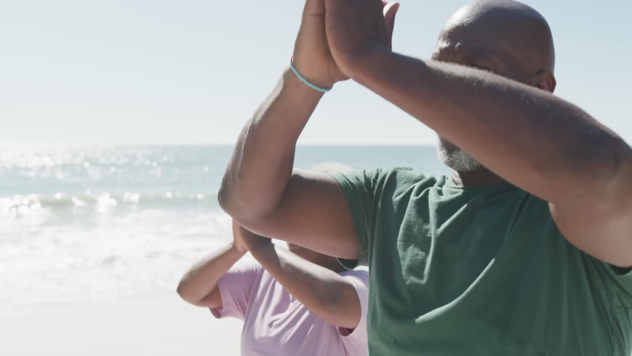 Happy senior african american couple doing yoga and meditating at beach, in slow motion