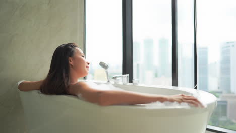 while relaxing in a large resort-style bathtub, a young beautiful woman looks out on a modern skyline