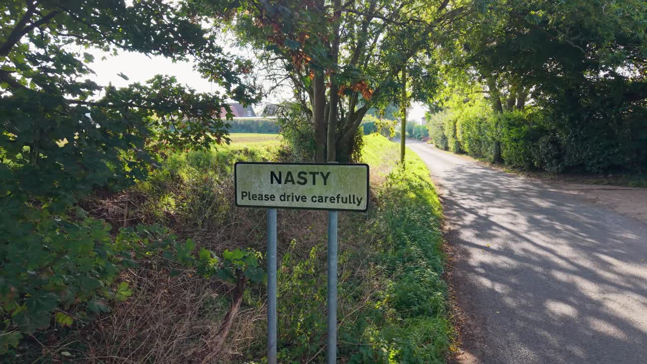 Aerial camera tracks right across the Nasty, Hertfordshire hamlet sign, ending at an angle. Shafts of light filter through surrounding trees, creating a blend of rural charm, atmosphere, and metaphor