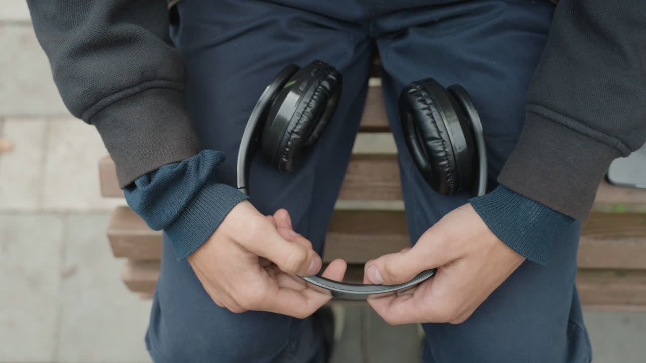 hombre sentado en un banco sosteniendo unas gafas y unos auriculares sobre el regazo, con un jersey de punto de varias capas y pantalones azules, ajustando las monturas con los dedos en una pausa urbana tranquila, primer plano espontáneo de las manos y el accesorio, en tonos apagados