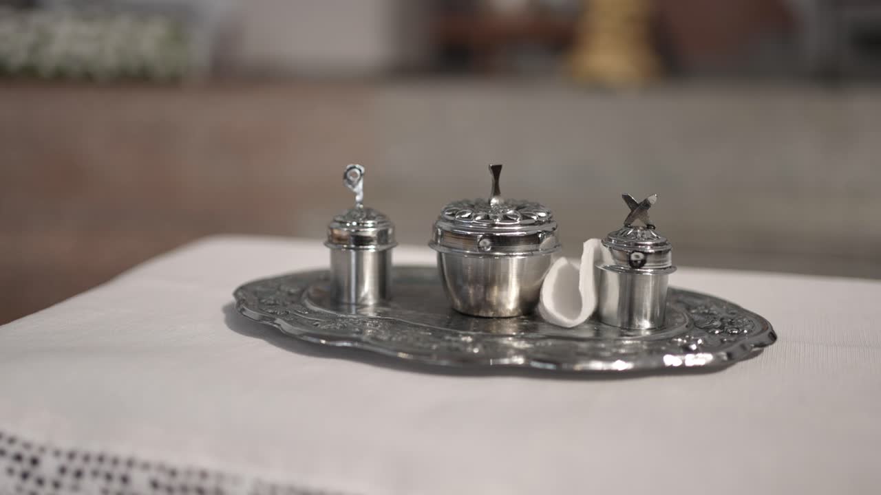 Close up of silver vessels and tray used in Catholic baptism ceremony placed on white altar cloth
