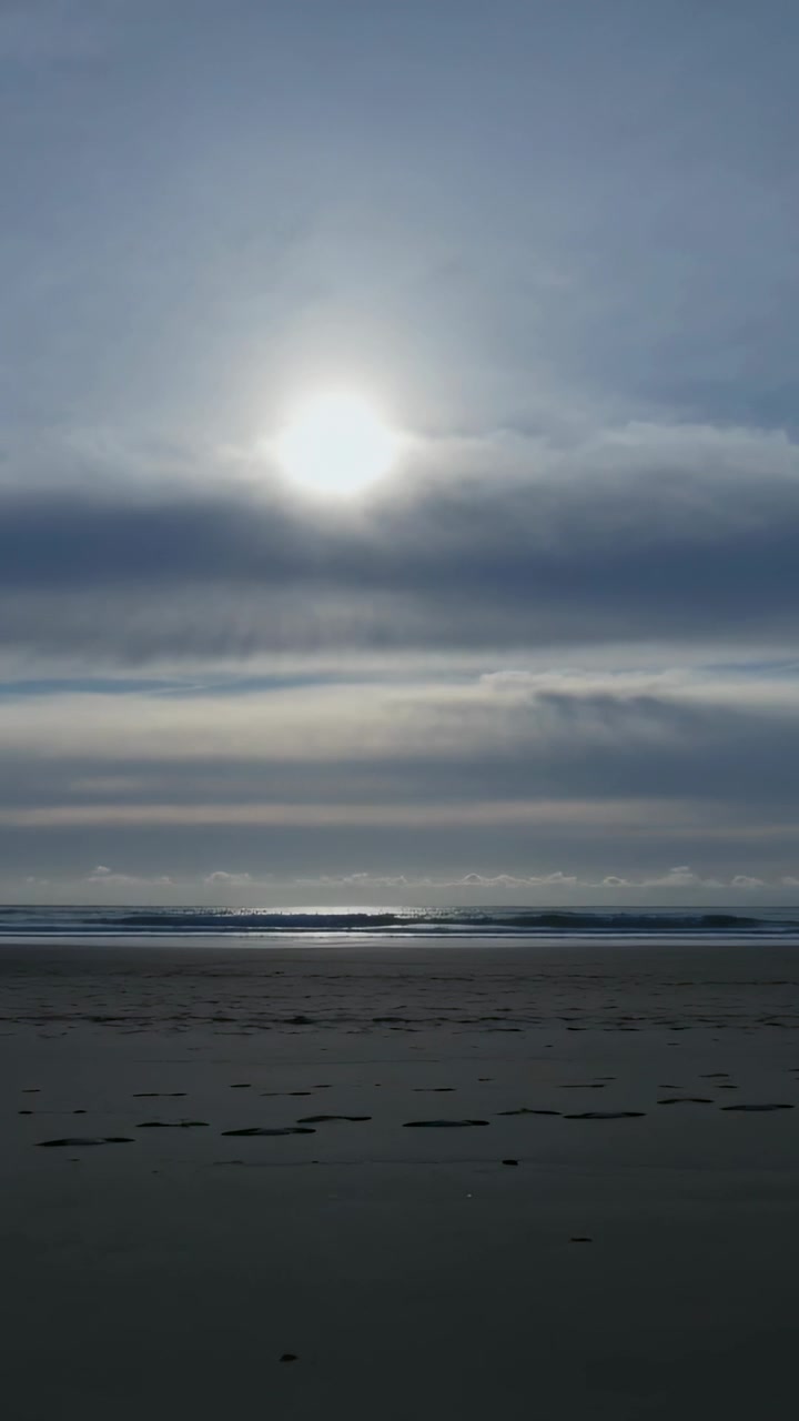 Vertical video: Shining sun piercing cloud band over wet sand, causing rippling water reflections