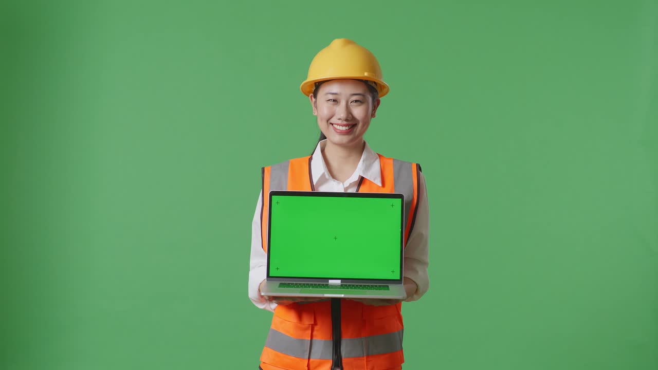 안전 헬을 입은 아시아 여성 엔지니어가 미소 짓고 녹색 스크린 백그라운드 스튜디오에 서 있는 동안 카메라에 녹색 화면 노트북을 보여주고 있습니다.