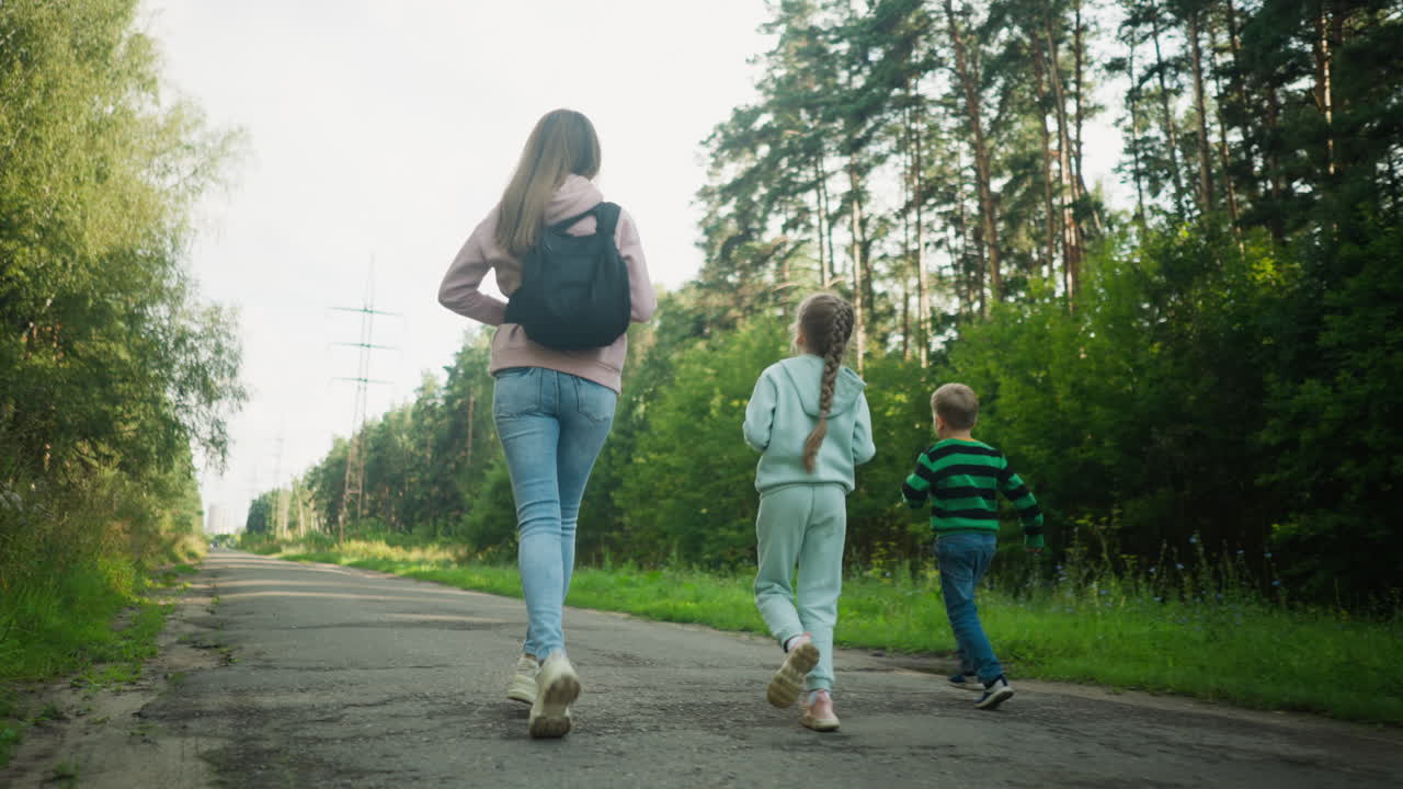 Rear view of young siblings joyfully running beside older sister on paved forest road, surrounded by tall trees and peaceful greenery, capturing energetic family bonding in nature on sunny day