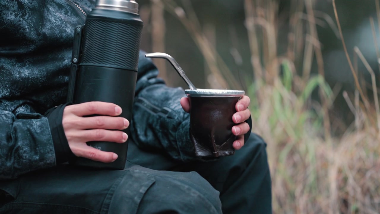 Una persona versa acqua calda da un thermos in una tazza da mate, seduta all'aperto in un bosco. Ideale per temi di lifestyle o avventura all'aria aperta.
