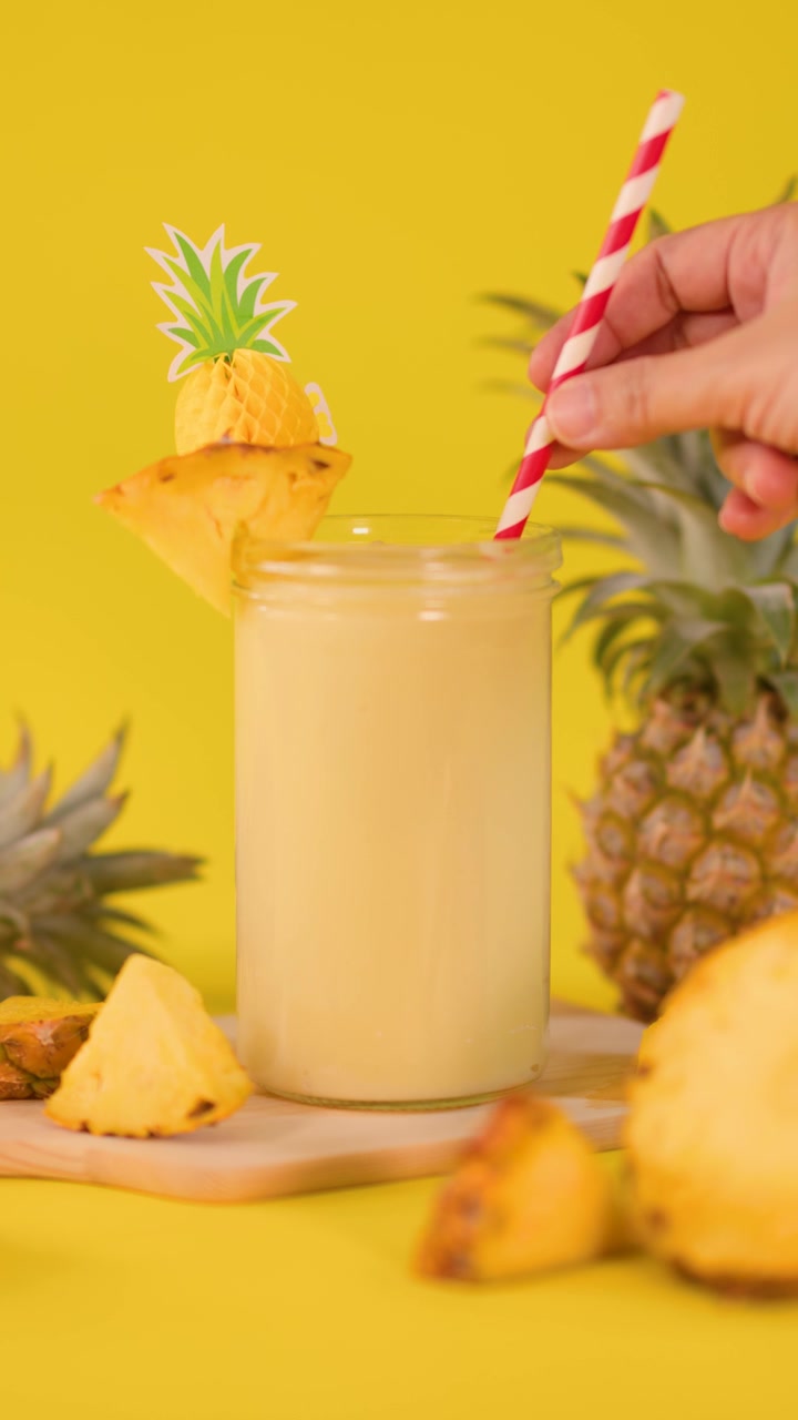 Una mano inserisce una cannuccia a strisce in un frullato cremoso all'ananas, circondato da frutta fresca, su uno sfondo giallo brillante.