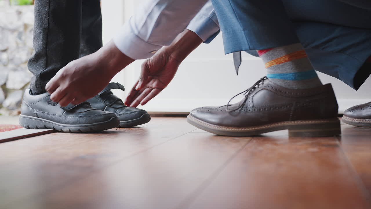 Close up of father wearing shirt and tie, brogue shoes and striped socks kneeling down on one knee to fasten the straps on his son&rsquo;s shoes before school, low section, close up of feet