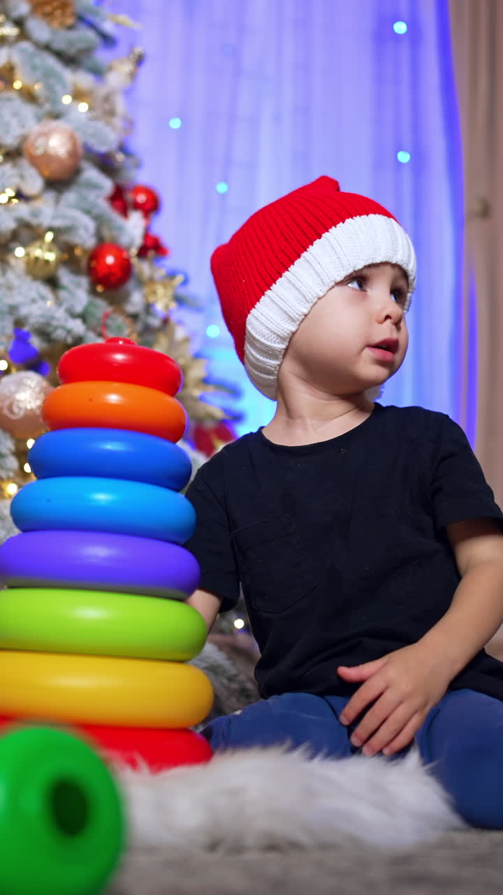 Adorable Caucasian toddler in red cap sitting near the Christmas tree. Kid pushes the toy pyramid falling on the floor. Low angle view. Vertical video
