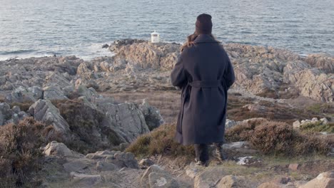 woman watching lighthouse at sunset at rocky cliff while her coat waves in the wind