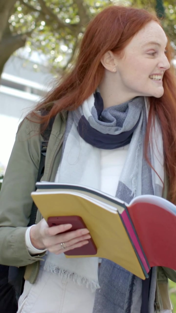 Video verticale: Donna che cammina e prova le battute da un quaderno al parco, guardando a destra, tenendo il telefono.