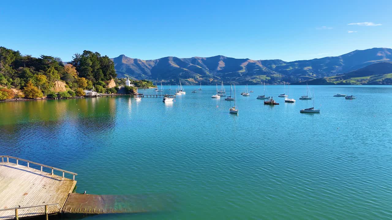 Calm waters with anchored boats under clear skies in Akaroa, New Zealand. Scenic views of hills and tranquil harbor setting