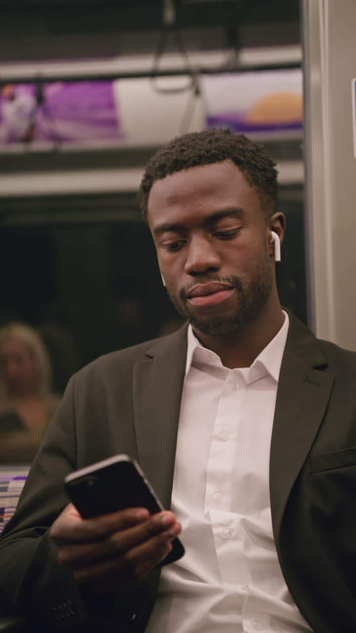 Vertical Video Of Young Businessman In Suit Commuting To Work Sitting In London Underground Tube Train Wearing Wireless Earbuds To Stream From Mobile Phone Shot In Real Time