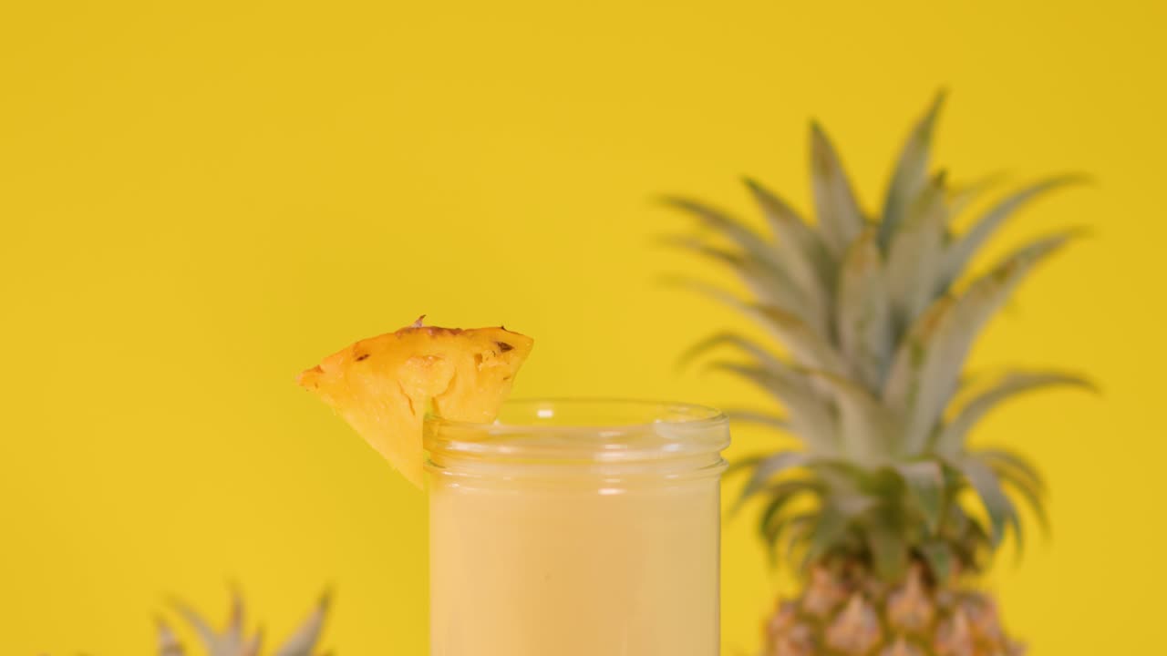 Hand places pineapple-shaped stirrer in creamy smoothie beside pineapple slice, bright yellow backdrop, static shot