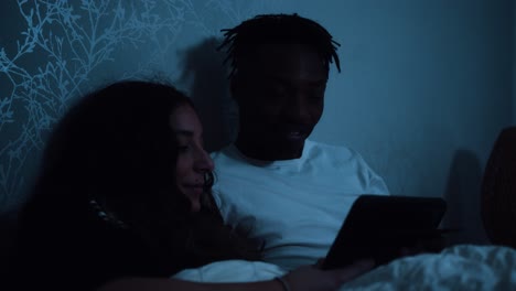 una pareja joven sentada en la cama por la noche viendo un dispositivo de tableta