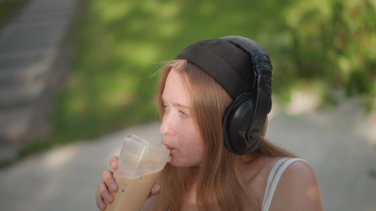 young woman sipping iced coffee on sidewalk, wearing headphones and beanie, casual tank, golden sunlight and bokeh park path, thoughtful expression, student break and freelance writer vibe, candid