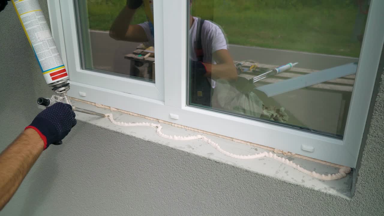Nærbillede af arbejderhænder i beskyttelseshandsker, der påfører polyurethan byggeskum på en vindueskarm.