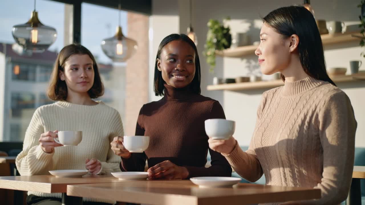 Drei Frauen in Strickpullovern nippen lächelnd an Kaffee aus weißen Tassen am Caféfenster.