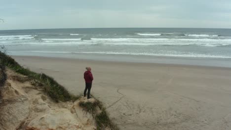 Un-Hombre-Se-Encuentra-Sobre-Una-Alta-Duna-De-Arena-Con-Vista-A-Una-Hermosa-Playa-Y-Vista-Al-Mar-En-Oregon