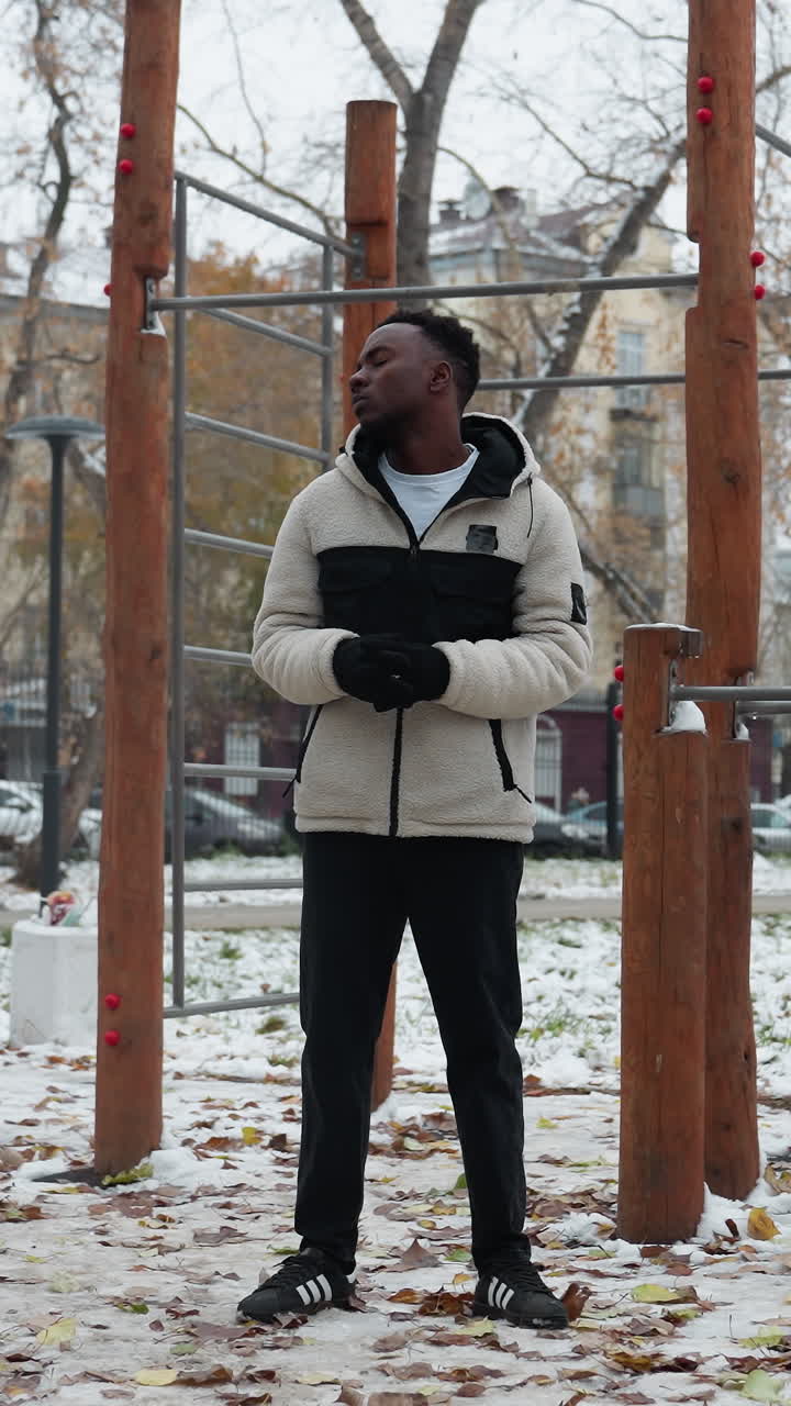 joven adulto realizando torsión del cuello al aire libre en chaqueta de invierno, manos unidas, de pie en medio de equipos de entrenamiento en un parque urbano, suelo cubierto de nieve y árboles desnudos en el fondo