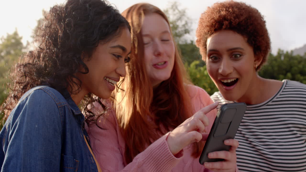 Hanging out outdoors, female friends smiling and pointing at smartphone, enjoying time together