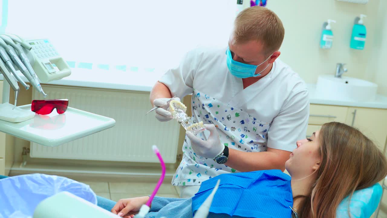 Una niña en el dentista examina el modelo de mandíbula. Un médico con mascarilla y guantes de goma explica el procedimiento de tratamiento.