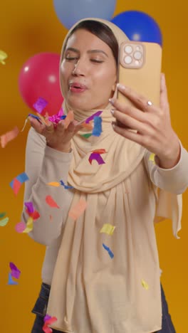 Vertikales-Videostudio-Porträt-Einer-Frau,-Die-Ein-Selfie-Mit-Hijab-Macht-Und-Geburtstag-Feiert-Und-Von-Luftballons-Umgebenes-Papierkonfetti-Bläst