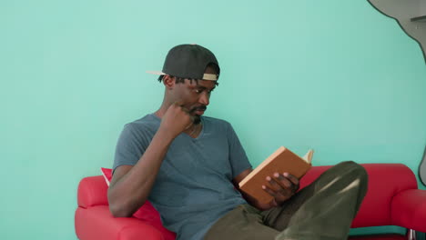 Literature lover in casual shirt and cap sits on red couch with crossed leg, holding open book and reading intently with focused expression in bright indoor room with turquoise wall and bookshelf