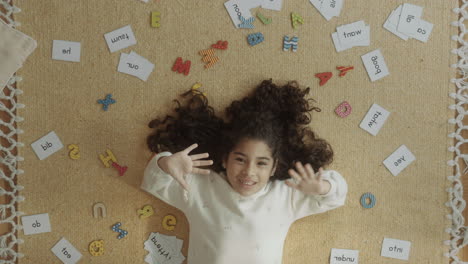 jolly little girl lying among word cards and waving at camera