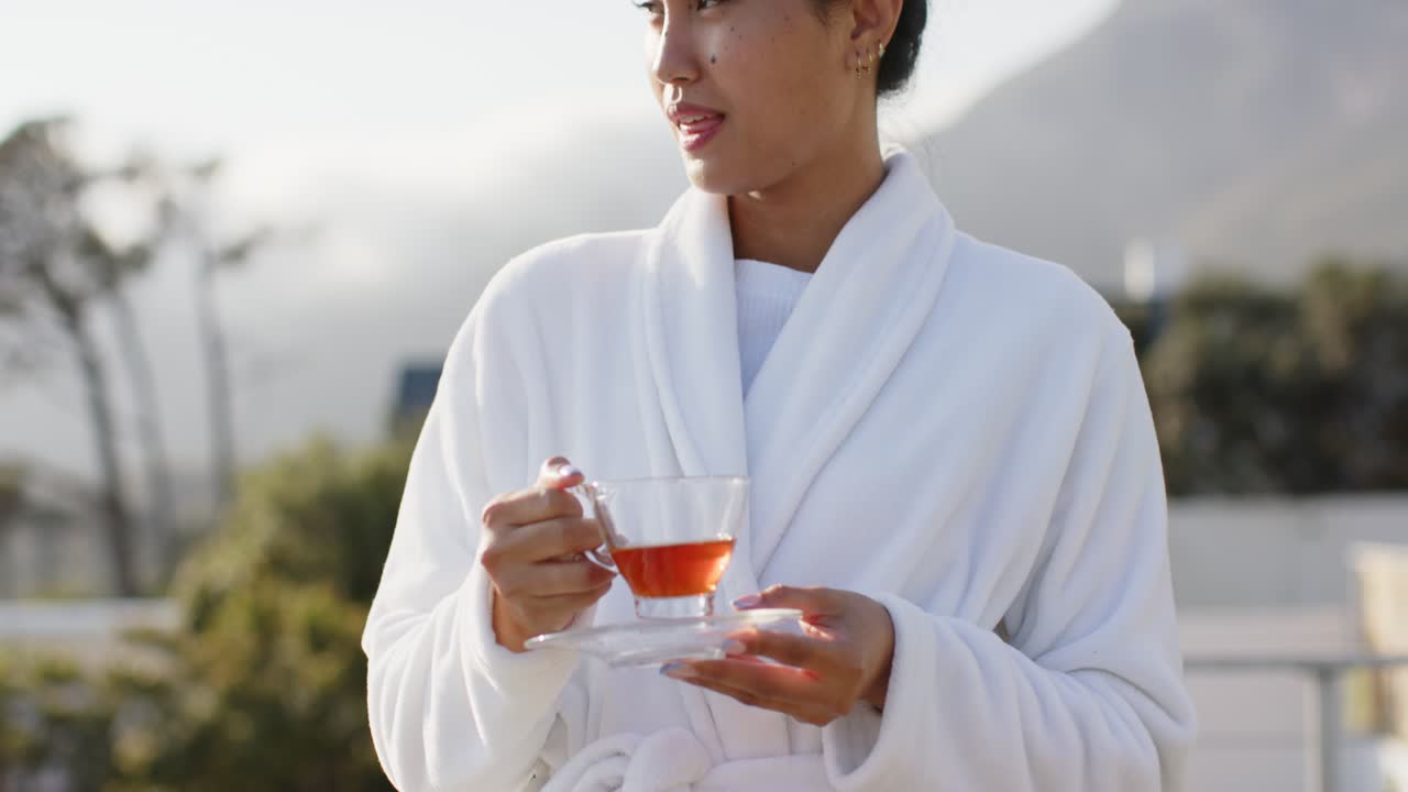 Donna asiatica ispirata dalla luce del mattino che solleva una tazza sorseggiando tè, appoggiando la tazza sul piattino, ammirando la montagna
