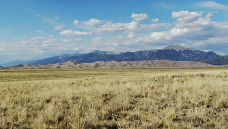 Aerial-cinematic-drone-late-summer-opening-view-entrance-of-The-Great-Sand-Dunes-National-Park-Colorado-Rocky-mountain-14er-peaks-crisp-golden-yellow-tall-grass-blue-sky-slide-to-the-left-movement