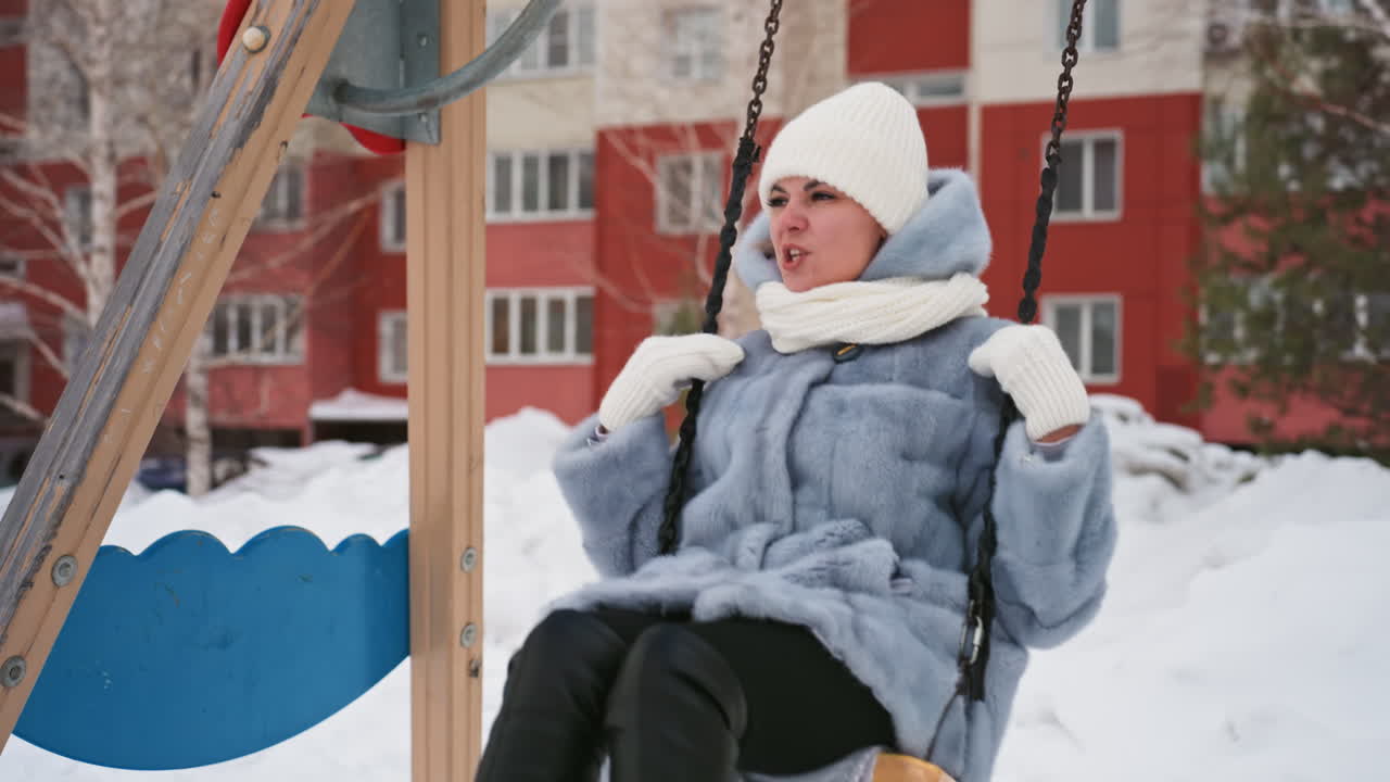 Mamma sull'altalena in un parco giochi innevato, avvolta in un cappotto blu e cappello bianco, sorride dolcemente, scena urbana invernale pacifica vicino ai palazzi, mani che stringono le catene, impronte nella neve, atmosfera accogliente e contemplativa