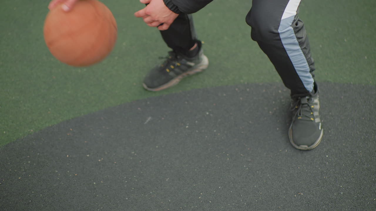 Pebasket yang sedang latihan menggiring bola dengan posisi merendah di lapangan luar ruangan, melakukan rangkaian dribel rendah di permukaan karet bertekstur, sikap lebar yang stabil, sol sepatu yang mencengkeram, tangan mengendalikan bola, persiapan crossover