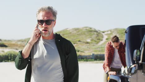 stressed caucasian gay male couple standing by broken down car, talking on smartphone at the beach